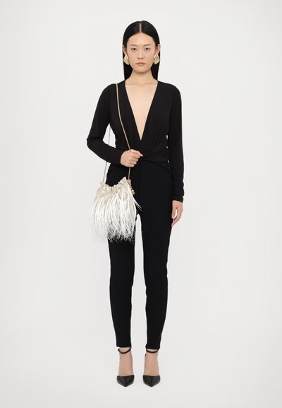 Black long-sleeve top with deep V-neck, black pants, and a light handbag with white fringe detail. Model wearing black heels and large earrings.