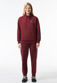Burgunderfarbener Hoodie und passende Jogginghosen aus weicher Stoffqualität, mit einer Fronttasche und Kapuze mit Kordelzug. Mit einem grünen Logo akzentuiert.