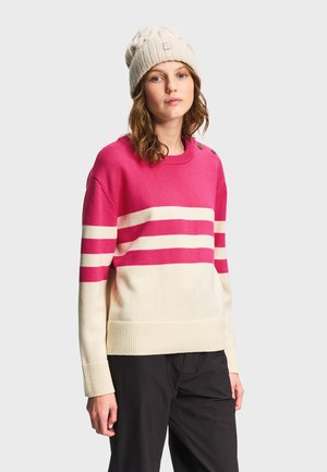 Jeune femme portant un bonnet en laine blanc et un pull rayé rose et blanc, debout devant un fond gris clair.