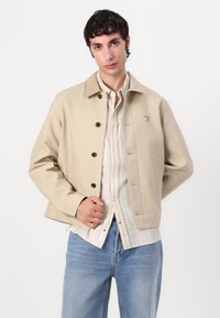 Beige uformell jakke med spiss krage, knappelukking og to frontlommer. Brukt over en lysstripete skjorte. Har subtile logo detaljer.