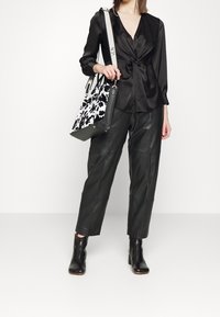 Femme portant un chemisier en satin noir, un pantalon en cuir noir, des bottines, tenant un sac fourre-tout à motif noir et blanc avec une sangle blanche.
