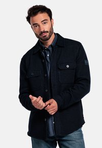 Man met kort zwart haar en baard, gekleed in een donker button-up shirtjack met klepzakken, terwijl hij de manchette vastknopt en tegen een witte achtergrond staat.