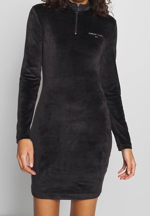 Schwarzes Velourskleid mit langen Ärmeln, Halbzip-Kragen und kleinem Tommy Jeans-Logo auf der Brust, getragen von einer Person mit lockigem Haar.