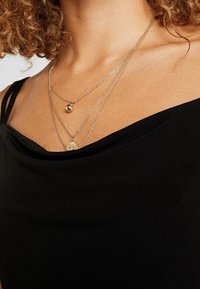 Femme portant des colliers en or superposés avec des pendentifs ronds et ovales sur un top noir à épaules dénudées, montrant des cheveux bouclés et un cou nu.
