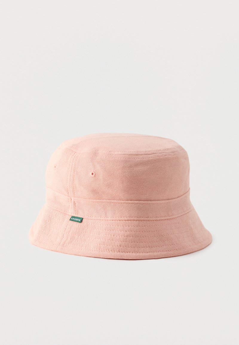 Hellrosa Stoff-Bucket-Hut mit kleinem grünem Lacoste-Logo-Label an der Seite, weicher Textur und genähtem Rand auf einfarbigem Hintergrund.