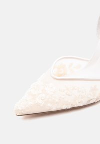 Chaussure blanche à bout pointu avec dentelle florale et détails en sequins sur un fond uni, montrant la face avant et le côté intérieur.
