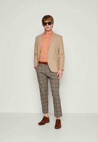 Homem vestido com um blazer bege, camisa laranja de botão, calças em padrão xadrez, cinto e sapatos castanhos, e óculos escuros pretos, de pé contra um fundo simples.