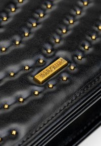 Czarny skórzany portfel z pikowaną fakturą, złotymi ćwiekami oraz prostokątną złotą tabliczką z wytłoczonym napisem.