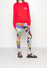 Sudadera roja con el logo "love moschino", combinada con leggings de patrón abstracto multicolor. El calzado presenta sandalias negras con marca.