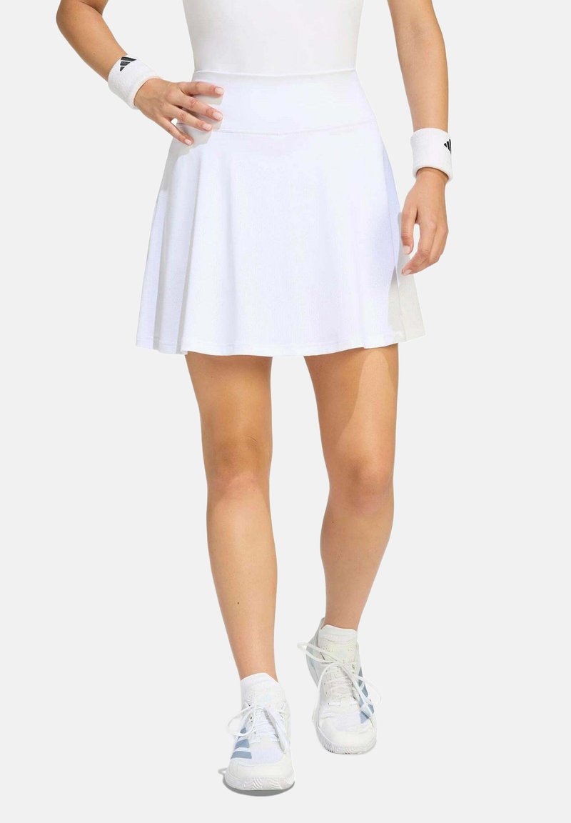 Femme portant une jupe de sport blanche, des chaussures de sport blanches et des bracelets de poignet blancs, marchant avec une main sur la hanche.