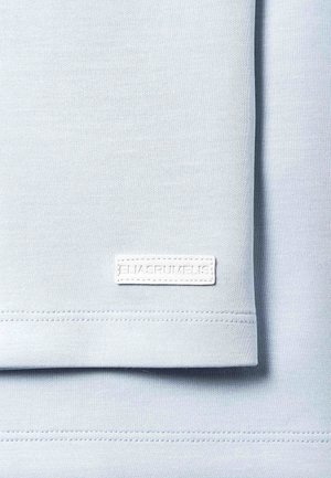 Vaaleansininen kangas taitettuna, jossa pieni valkoinen suorakaiteen muotoinen etiketti pohjareunassa, jossa on kohokuvioitu teksti "ELIASRUMELIS".