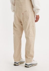 Mono beige a rayas con un corte holgado. Cuenta con grandes bolsillos traseros y tirantes ajustables. Combinado con zapatillas blancas.