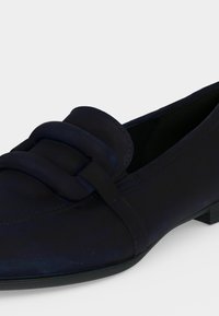 Mocassin en cuir noir à enfiler avec bout arrondi et bride matelassée décorative sur le dessus, petit talon empilé, sur fond blanc uni.