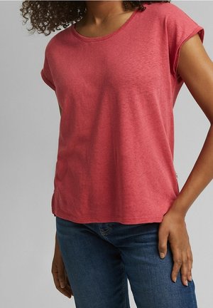 Frau trägt ein kurzärmeliges, korallenrotes T-Shirt und blaue Jeans, steht mit einer Hand auf der Hüfte.