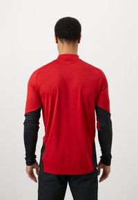 Röd idrottströja med långa ärmar som har svarta mönstrade ärmar, ståkrage och en figurnära design. Slätt, lättviktigt tyg.