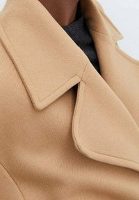 Beżowy płaszcz z wełny z szerokim, klapowym kołnierzem i strukturalną tkaniną. Zdjęcie z bliska podkreśla szczegóły kołnierza i gładkie wykończenie.
