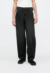 Άτομο που φορά φαρδιά ξεθωριασμένα μαύρα τζιν, λευκό τοπ και μαύρα αθλητικά παπούτσια, στέκεται σε απλό φωτεινό φόντο.