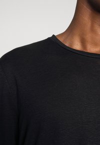 Gros plan sur le cou et l'épaule d'une personne portant un t-shirt noir uni à col rond sur un fond gris clair.