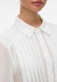 Camicia bianca con bottoni, caratterizzata da pieghe verticali, un colletto a punta e un tessuto leggero e trasparente. Accentuata da bottoni classici.