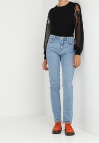 Zwarte top met doorzichtige, geborduurde mouwen, lichtblauwe fitted jeans en oranje schoenen. Gladde stof en slanke silhouet.
