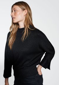 Haut en tissu noir pailleté avec un col montant et des manches trois-quarts ; il présente une coupe décontractée et un ourlet côtelé.