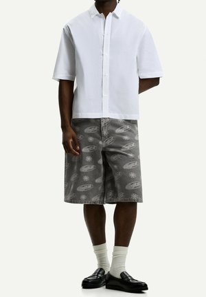 Camicia bianca a maniche corte con bottoni abbinata a pantaloni corti grigi con motivo, caratterizzati da una grafica "digitale", completata da mocassini neri e calzini bianchi.