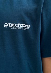 Tamno plava pamučna majica s bijelim "project core" logotipom i tekstom, s okruglim izrezom i kratkim rukavima. Glatka tekstura.