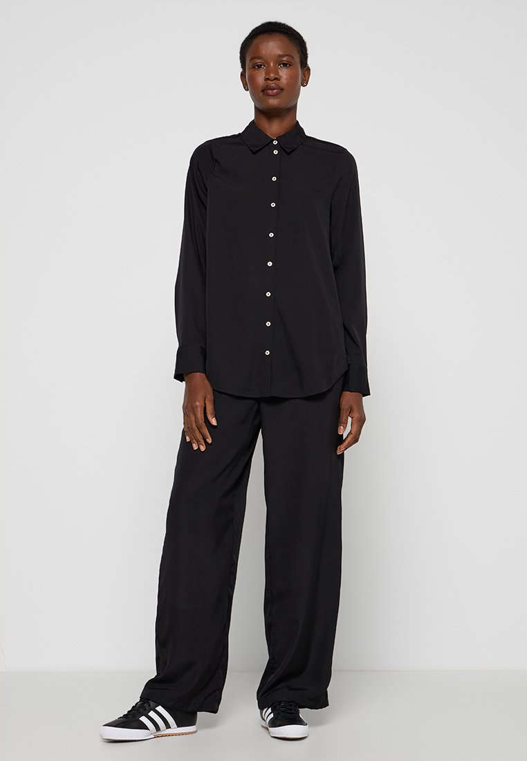 Zwarte langemouwen shirt met knoopsluiting, kraag en een losse pasvorm, gecombineerd met losse zwarte broek en zwart-witte sneakers.