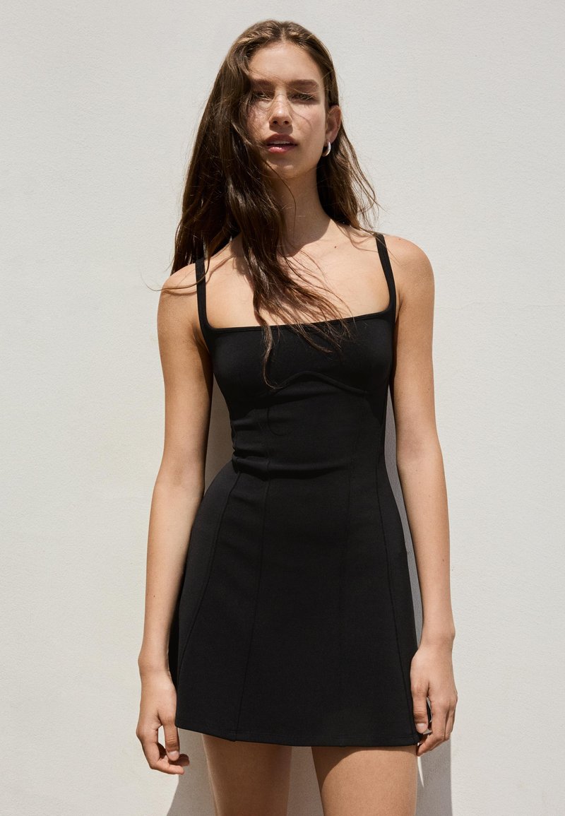 Femme aux longs cheveux bruns portant une robe noire moulante sans manches, debout contre un mur clair uni.