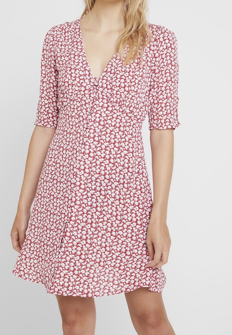 Červené květinové šaty s krátkými rukávy, výstřihem do V a rozšířenou siluetou. Tkanina má po celém povrchu drobný bílý květinový vzor.