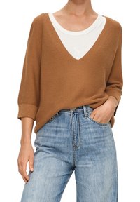 Brun stickad tröja med bred V-ringning lagd över en vit skjorta. Tillverkad med ljusblå högmidjade jeans. Avslappnad design.