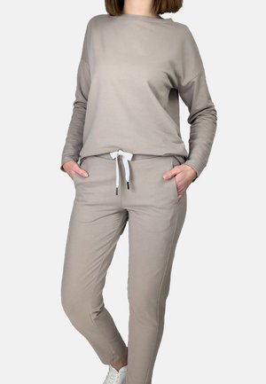 Person wearing a beige long-sleeve sweatshirt and matching jogger pants with a white drawstring, hands in pockets, white sneakers visible.