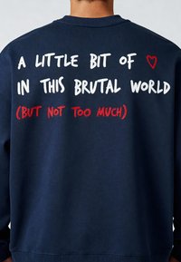 Tumši zils džemperis ar izsistiem burtiem uz muguras: "NEDAUDZ ♥ ŠAJĀ BRUTĀLĀ PASAULĒ (BET NE PĀRMĒRĀ)." Mīksta tekstūra, apaļas kakla dizains.
