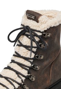 Stivale da trekking in pelle marrone con fodera in pile crema, lacci neri e occhielli metallici. Presenta un patch con il logo nella parte superiore.