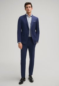 Herrenanzug in Marineblau aus glattem Stoff, mit einem Blazer mit zwei Knöpfen und schlankem Schnitt. Kombiniert mit einem hellblauen Hemd und schwarzen Schuhen.