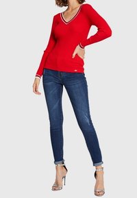 Punainen ribbineulottu neulepusero V-pääntiellä ja valkoisilla raitakuvioilla pääntiessä ja hihansuuissa, yhdistetty tummansinisiin kapeisiin farkkuihin ja hopeisiin korkokenkiin.