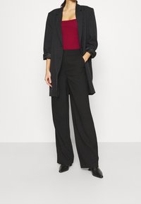Donna che indossa un lungo blazer nero, pantaloni neri a gamba larga, scarpe nere punti e un top senza maniche color bordeaux su uno sfondo semplice.