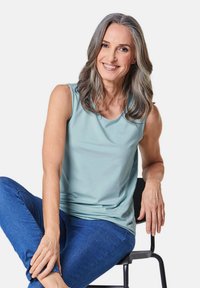 Top senza maniche di colore azzurro chiaro realizzato in un tessuto liscio, abbinato a jeans blu. Modella seduta su una sedia nera, in una postura rilassata.
