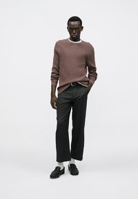 Brun ribstrikket sweater over en hvid skjorte, kombineret med grå bukser med vide ben. Sorte loafers og hvide sokker fuldender outfittet.