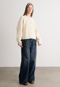 Cárdigan de punto cable crema con cuello redondo y botones frontales, combinado con unos vaqueros oscuros holgados y calzado negro.