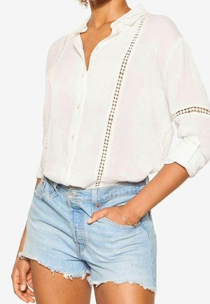 Femme portant un chemisier blanc boutonné avec des bordures en dentelle et des manches retroussées, associé à un short en denim bleu clair effiloché.