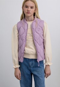 Gilet matelassé lavande avec design matelassé, fermeture éclair avant et détail logo, porté sur un pull crème avec un motif graphique, associé à un jean bleu.