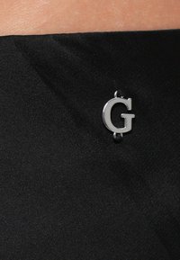 Črna tkanina z gladko teksturo, na kateri je srebrni logotip "G" za poudarek. Logotip je pritrjen z majhnim sponkom.