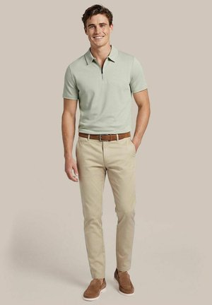 Junger Mann lächelnd, trägt ein hellgrünes kurzärmeliges Polo, beige Hose mit braunem Gürtel und braune Velours-Slipper, steht vor beigem Hintergrund.