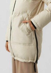 Beige pufferjack met een rits aan de voorkant, grote zijzak en zwarte accenten aan de manchetten. Heeft een zachte, gequiltte textuur.