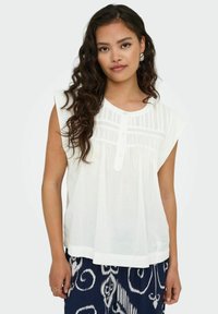 Blouse blanche sans manches avec un plastron à boutons et des plis verticaux à l'avant, faite d'un tissu léger. Associée à un pantalon foncé à motifs.