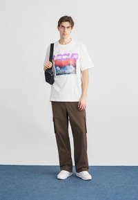 Bijela majica s ljubičasto-ružičastim grafičkim printom, smeđe cargo hlače i mala crna torbica preko tijela. Bijele tenisice upotpunjuju izgled.