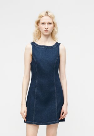 Lyshåret kvinde med bølget hår iført en ærmeløs mørkeblå denimkjole, stående mod en enkel lys grå baggrund.