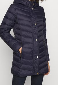 Granatowa kurtka puchowa z kapturem, z poziomymi przeszyciami, z przodu na zatrzaski, bocznymi kieszeniami na zamek i eleganckim, lekkim designem.