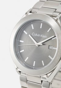 Reloj de acero inoxidable plateado con un dial circular cepillado en gris, marcadores de horas blancos y display de fecha a las 6 en punto. Pulsera de eslabones de metal.